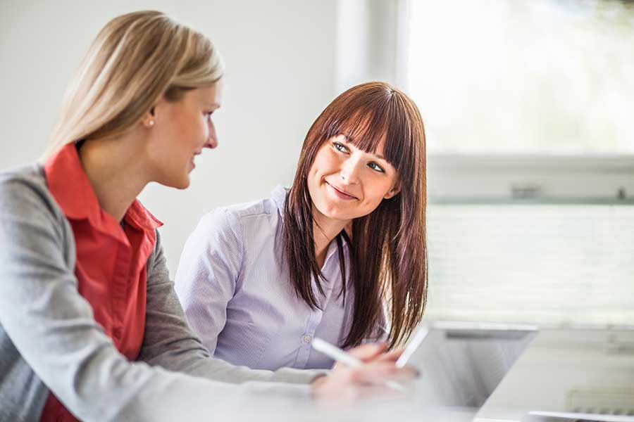 Two German women talking and smiling at a desk with documents.