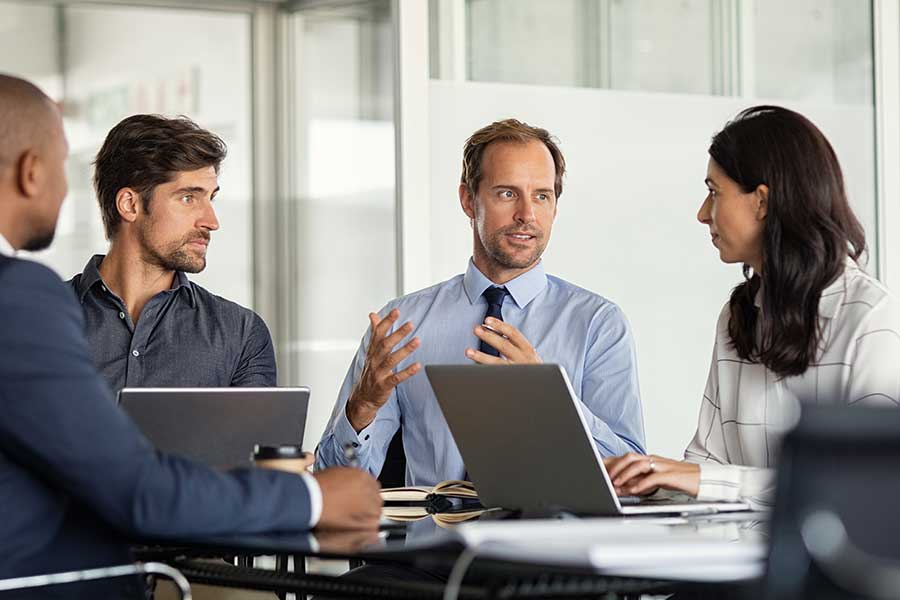 Three men and one woman at a meeting with laptops open. Man gesturing.