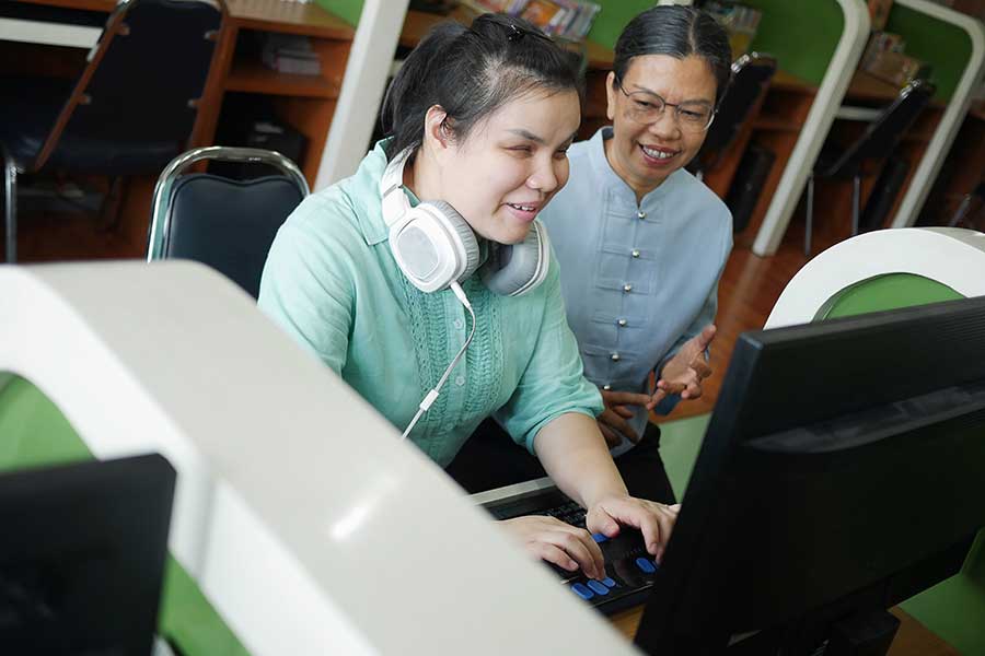 Asian young blind person woman with headphone using computer with braille display assistive device discussing with senior colleague woman in workplace.